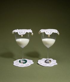 two wine glasses filled with white liquid and garnished with green beads, sitting on lace doily