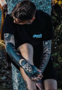 a man with tattoos on his arm sitting next to a tree