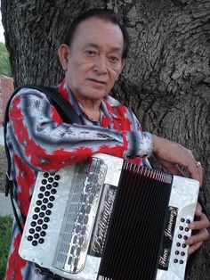 an old man is playing the accordion in front of a tree with his arms crossed