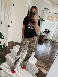 a woman taking a selfie in the mirror with her cell phone while wearing camo pants and red shoes