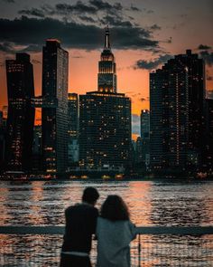 two people sitting on a bench in front of the city skyline at sunset or dawn