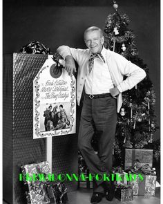 a man standing next to a christmas tree with presents in front of him and a sign that says merry christmas