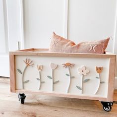 a decorative wooden box with flowers on the sides and wheels, sitting on a wood floor in front of a white door