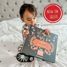 a small child is sitting on a bed and holding a book that says now on sale