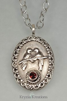 This Victorian-inspired love birds necklace aims to celebrate the shared love and devotion between two people in timeless style. Handcrafted from oxidized sterling silver, a decorative crown border encircles an oval pendant surrounding two devoted love birds sitting on a branch atop a small faceted garnet. See and shop this one-of-a-kind necklace at www.KryziaKreationsStudio.com