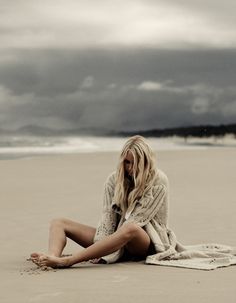 a woman sitting on top of a sandy beach next to the ocean under a cloudy sky