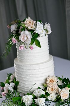 a white wedding cake with pink and white flowers on the top is surrounded by greenery