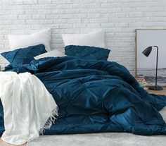 a bed with blue comforter and pillows in front of a white brick wall next to a lamp