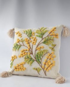 a white pillow with yellow flowers and tassels on the front, sitting against a gray background