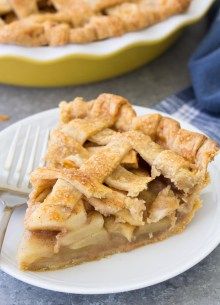 a slice of apple pie on a white plate with a fork and bowl in the background