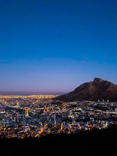 the city lights are lit up at night as seen from atop a hill with mountains in the background