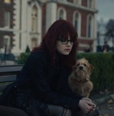 a woman with red hair sitting on a bench next to a small dog and looking at the camera