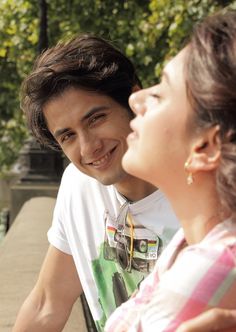 a young man and woman sitting next to each other on a park bench smiling at each other