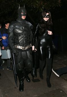 two people dressed up as batman and catwoman walking down the street at night time