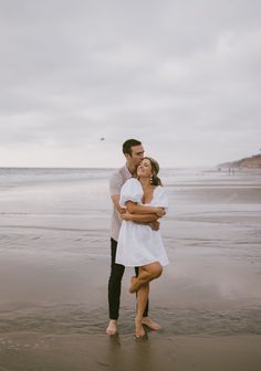 a man and woman hug on the beach while standing next to each other in front of the ocean