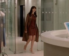 a woman in a dress and trench coat is walking through a glass walled room with a laptop