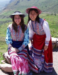 two women in colorful dresses and hats posing for the camera on top of a hill