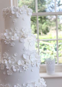 a three tiered white wedding cake with flowers on the top and bottom, sitting in front of a window