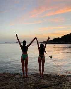 two women in bikinis standing on the beach with their arms up to the sky