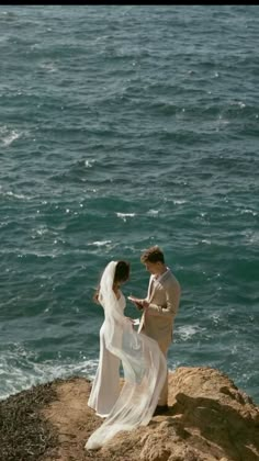 a bride and groom are standing on the rocks by the water