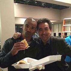 two men sitting at a table with food in their hands and one holding a sandwich