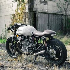 a motorcycle parked in front of a building on the side of a dirt road with weeds growing around it