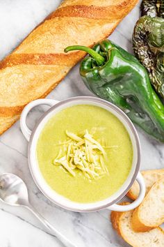 a bowl of broccoli soup next to bread and green peppers on a marble surface