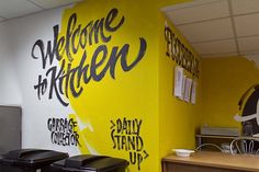 there are two black trash cans in front of the yellow wall that says welcome to kitchen