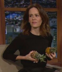 a woman sitting on a couch holding pineapples and looking at the camera while wearing a black dress