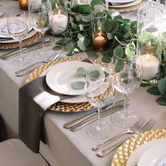 the table is set with white and gold plates, silverware, candles and greenery