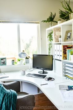 a white desk topped with a computer monitor next to a window filled with green plants