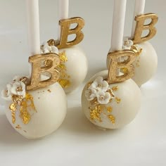 three white cake pops with gold letters and flowers on them, sitting next to each other