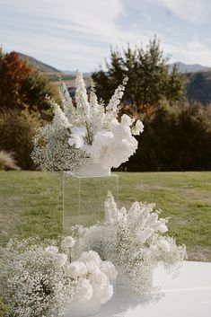 white flowers in vases sitting on top of a table with grass and mountains in the background