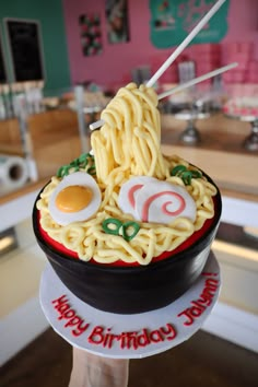 a person holding up a cake with noodles and eggs on it in the shape of a ramen bowl