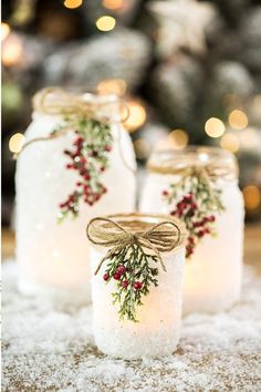 three mason jars decorated with holly and berries are sitting on snow in front of a christmas tree