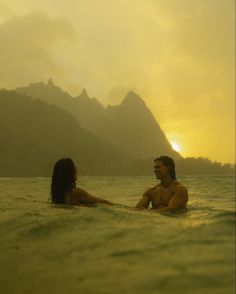 two people are in the water with mountains in the backgrouds and one person is sitting on his surfboard