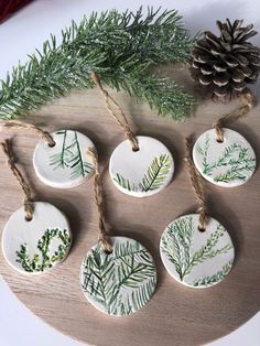 six ceramic ornaments on a wooden board with pine cones and twine tied to them