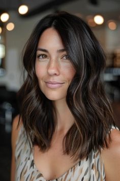 Woman with medium-length wavy brunette hair, wearing a patterned top, smiling softly indoors. Chocolate Brunette Hair With Highlights, Brunette Hair Ash, Dark Medium Hair With Highlights, Brunette All Over Hair Color, Ombré On Black Hair, Chocolate Medium Length Hair, Dark Brown Hair Chocolate Highlights, Brown Hair Balayage Dark, Brunette No Highlights