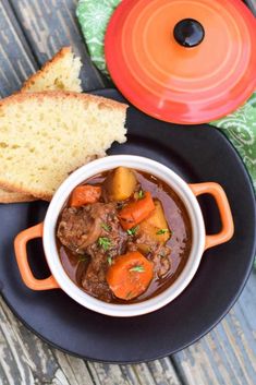 a black plate topped with a bowl of stew next to a piece of bread on top of a wooden table