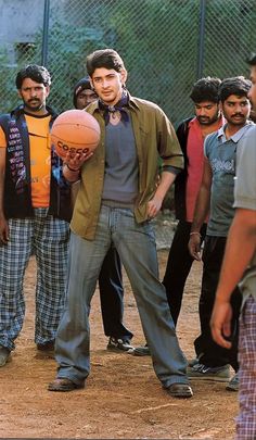 a young man holding a basketball in front of other men on a dirt ground with a chain link fence behind him