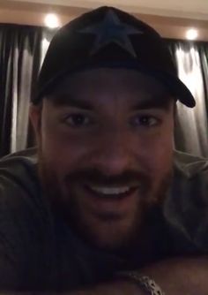 a man with a beard wearing a baseball cap and smiling at the camera while sitting in front of a curtain