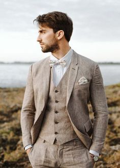 a man wearing a suit and bow tie standing in front of the ocean with his hands in his pockets