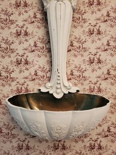 a white bowl with a metal spoon in front of a wallpapered wall and a mirror above it