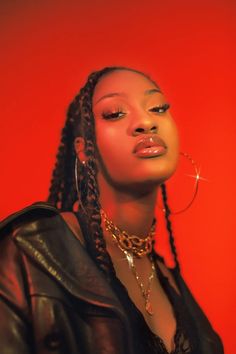 a woman with braids wearing a black leather jacket and gold hoop earrings, against a red background