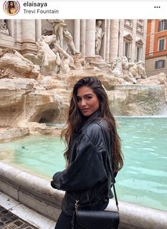 a woman standing in front of a fountain with a purse on her shoulder and looking at the camera