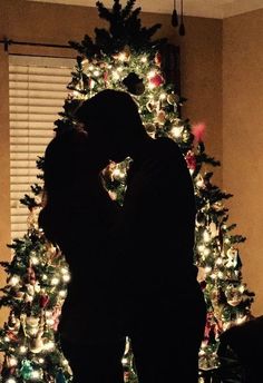 a man and woman kissing in front of a christmas tree with lights all around it