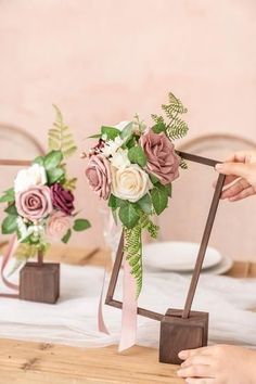 two vases with flowers and greenery are on a table, one being held up by a woman's hand