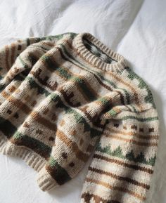 a sweater laying on top of a white bed next to a pillow with an orange, green and brown striped design