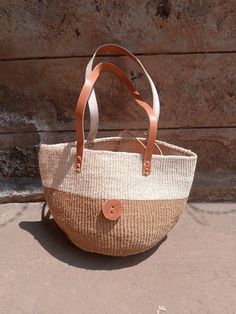 "Natural sisal kiondo bag with stitched leather straps. Ideal for the beach 🏖️, with a casual wear and a perfect accessory for an evening out with friends. This unique tote bag is handmade by a group of ladies in a remote rural village in Machakos, Kenya. DIMENSIONS Bottom width: 10\"/25.5cm Top width: 14\"/35.56cm Depth: 10\"/25.5cm Straps: 15\"/38cm SHIPPING All orders ship via DHL Express (3-5) days delivery time." Jute Bucket Bag For Market, Natural Bucket Bag With Braided Handles For Market, Market Basket Straw Bag With Leather Handles, Woven Leather Bucket Shoulder Bag, Handwoven Jute Bucket Bag For Daily Use, Market Basket Shoulder Bag With Leather Handles, Daily Use Basket Bucket Bag With Leather Handles, Natural Color Basket-shaped Bucket Bag With Adjustable Strap, Daily Handwoven Jute Bucket Bag