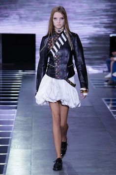 a woman walking down a runway wearing a black jacket and white skirt